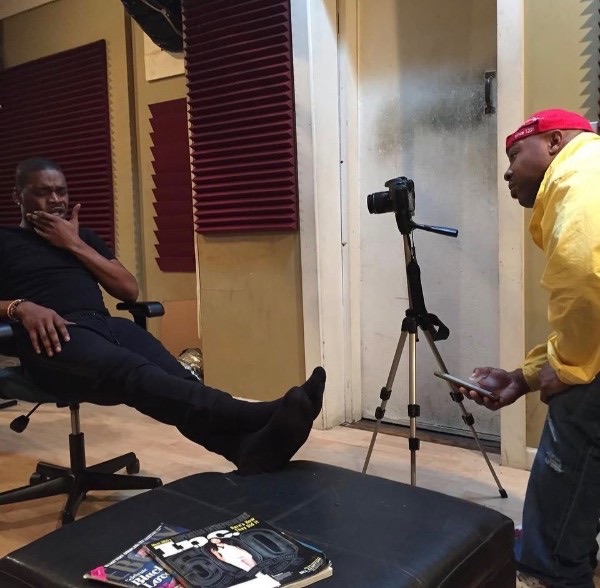 A camera on a tripod between a seated subject and a photographer in a recording studio