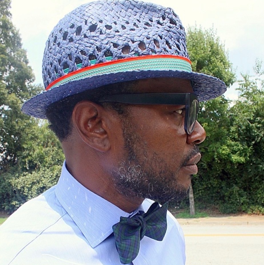 Profile portrait of a man wearing a woven hat, sunglasses, and bow tie